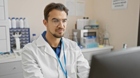 Foto de Un joven hispano con barba, gafas y bata de laboratorio, trabaja con atención en un laboratorio hospitalario. - Imagen libre de derechos