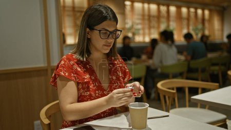 Téléchargez les photos : Belle femme hispanique verser du sucre dans le café du matin dans un café confortable, ses lunettes scintillant dans la lumière intérieure chaude - en image libre de droit