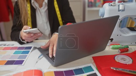 Téléchargez les photos : Une jeune femme caucasienne utilise un téléphone portable tout en travaillant avec du tissu et une machine à coudre dans un magasin de tailleurs. - en image libre de droit