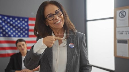 Confident woman with glasses pointing at her 'i voted' sticker in a room with another woman and american flag behind.
