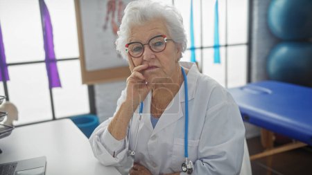 Foto de Una mujer mayor pensativa se sienta en una habitación de hospital, con una bata blanca, gafas y un estetoscopio.. - Imagen libre de derechos