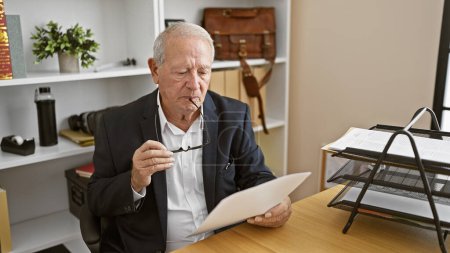 Téléchargez les photos : Homme âgé sérieux mais détendu, un professionnel prospère axé sur le progrès de l'entreprise, lisant attentivement un document sur son lieu de travail, exprimant sa confiance dans tous les cheveux gris - en image libre de droit