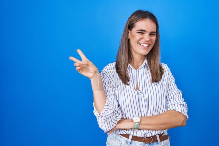 Téléchargez les photos : Jeune femme hispanique debout sur fond bleu souriant avec un sourire heureux clin d'oeil à la caméra faisant signe de victoire. numéro deux. - en image libre de droit