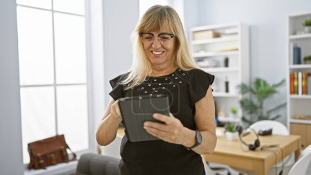 Photo for Confident middle age blonde woman worker, joyfully dominating the business scene, nailing her work on touchpad at the office. - Royalty Free Image