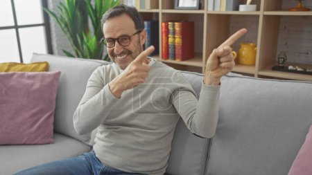 Foto de Sonriente hombre barbudo de mediana edad apuntando hacia los lados mientras está sentado en un sofá en una sala de estar moderna. - Imagen libre de derechos
