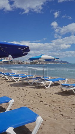 Téléchargez les photos : Une plage sereine avec chaises longues bleues et parasols sur un rivage sablonneux surplombant un paysage urbain côtier sous un ciel clair. - en image libre de droit