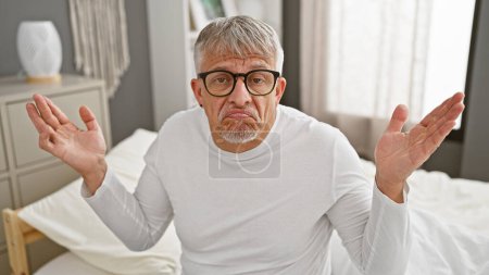 Verwirrter älterer Mann mit grauen Haaren und Brille, der auf einem Bett im Schlafzimmer sitzt, hebt fragend die Hände.