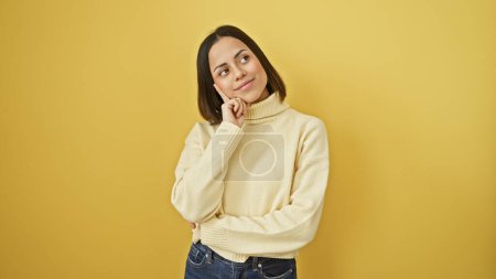 Téléchargez les photos : Jeune femme hispanique réfléchie avec une coiffure élégante posant sur un fond jaune isolé portant un pull décontracté. - en image libre de droit