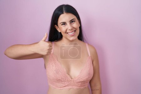 Foto de Mujer hispana joven con sujetador rosa haciendo un gesto feliz con la mano. aprobando la expresión mirando a la cámara mostrando éxito. - Imagen libre de derechos