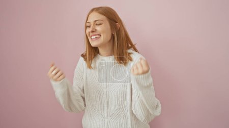 Photo for A cheerful young woman with redhead smiling joyfully in a white sweater against a pink background. - Royalty Free Image