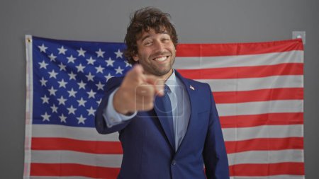Foto de Sonriente joven hispano de traje apuntando a la cámara con la bandera americana de fondo en un entorno de oficina. - Imagen libre de derechos