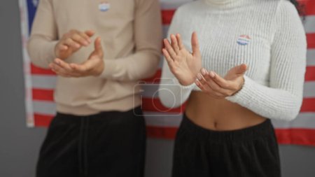 Man and woman with 'i voted' stickers applauding in front of an american flag in an indoor setting.