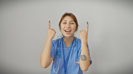 Foto de Una joven enfermera alegre con tatuajes apuntando hacia arriba, aislada sobre un fondo blanco, expresando positividad. - Imagen libre de derechos