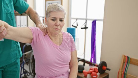 Eine Frau erhält Schulterphysiotherapie von einem männlichen Therapeuten in einem Klinikzimmer mit Trainingsgeräten.