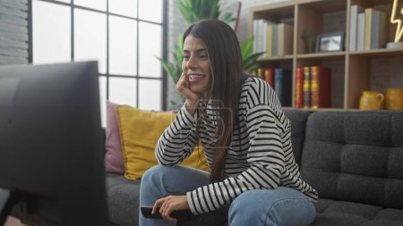 Téléchargez les photos : Une jeune femme hispanique sourit en regardant la télévision à la maison, main tenant à distance, dans un cadre confortable salon. - en image libre de droit