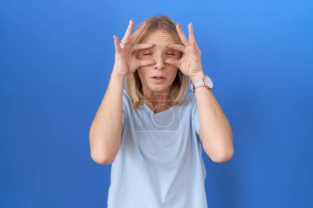Téléchargez les photos : Jeune femme caucasienne portant un t-shirt bleu décontracté essayant d'ouvrir les yeux avec les doigts, endormie et fatiguée pour la fatigue matinale - en image libre de droit