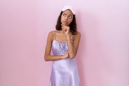 Photo for Young hispanic woman wearing sleep mask and nightgown thinking worried about a question, concerned and nervous with hand on chin - Royalty Free Image