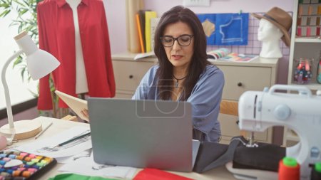Femme concentrée travaillant avec un ordinateur portable dans un atelier de tailleur, entourée de tissu, mannequin et machine à coudre.