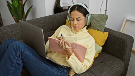 Téléchargez les photos : Hispanique femme étudiant avec écouteurs et ordinateur portable sur un canapé à l'intérieur - en image libre de droit