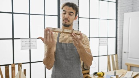 Téléchargez les photos : Bel homme hispanique inspectant le bois dans un atelier de menuiserie bien éclairé, transmettant l'artisanat et le souci du détail. - en image libre de droit