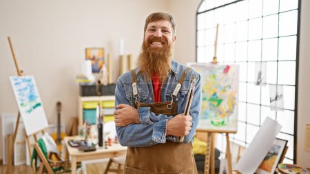 Téléchargez les photos : Un jeune rouquin confiant et souriant - un bel artiste dans un tablier les bras croisés, tenant un pinceau dans un atelier d'art pittoresque - en image libre de droit