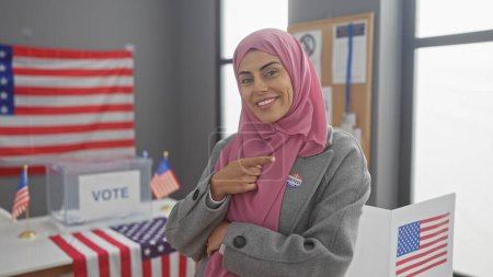 Téléchargez les photos : Une jeune femme confiante portant un hijab pointe son autocollant "j'ai voté" dans une salle d'université américaine ornée de drapeaux américains. - en image libre de droit