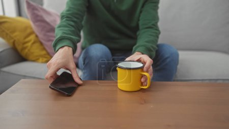 Foto de Un hombre casual se sienta con un teléfono inteligente y una taza en una mesa de café en un ambiente moderno de la sala de estar. - Imagen libre de derechos