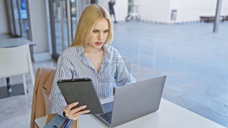 Téléchargez les photos : Une jeune femme concentrée travaille sur un ordinateur portable tout en tenant une tablette dans un cadre urbain extérieur moderne. - en image libre de droit