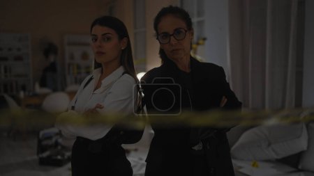 Two women standing with arms crossed at a crime scene indoors, exuding authority and professionalism.