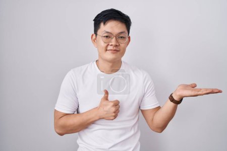 Foto de Joven asiático hombre de pie sobre blanco fondo mostrando palma mano y haciendo ok gesto con los pulgares hacia arriba, sonriendo feliz y alegre - Imagen libre de derechos