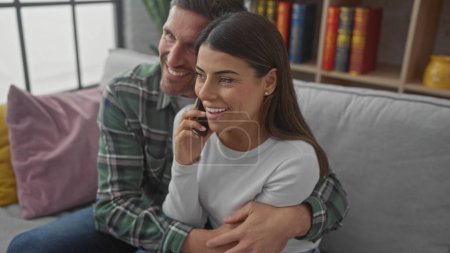 Foto de Una mujer sonriente en un teléfono abrazado por un hombre barbudo en un sofá en una acogedora sala de estar. - Imagen libre de derechos