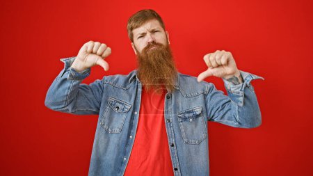 Cool, bearded redhead guy giving thumbs down  a negative sign of failure in casual lifestyle fashion, standing over an isolated red wall 