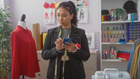 Foto de Una joven mujer hispana sastre en una chaqueta, sosteniendo un teléfono inteligente en un atelier colorido con maniquí y telas. - Imagen libre de derechos