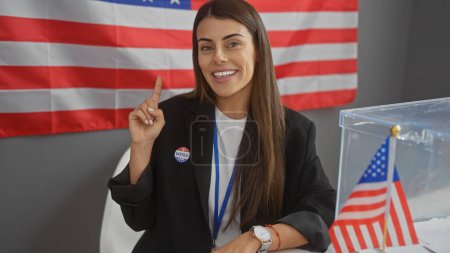Sonriente joven hispana con etiqueta de votante apuntando hacia arriba en una habitación con banderas y urnas americanas