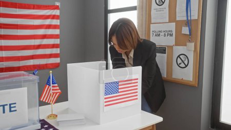 Foto de Una mujer afroamericana profesional votando en un centro electoral con una bandera americana en el fondo. - Imagen libre de derechos