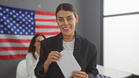 Smiling woman holding envelope with another woman and american flag in background at electoral center.