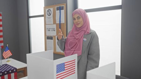A young woman with a hijab gives a thumbs-up in a voting booth with american flags.