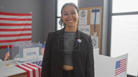 Lächelnde junge hispanische Frau mit "i voted" -Aufkleber im amerikanischen Wahllokal mit Fahnen