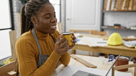 Téléchargez les photos : Femme afro-américaine avec des tresses profitant d'une pause café à son atelier de menuiserie à l'intérieur. - en image libre de droit