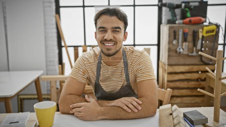 Foto de Hombre hispano sonriente con barba en taller de carpintería rodeado de herramientas - Imagen libre de derechos