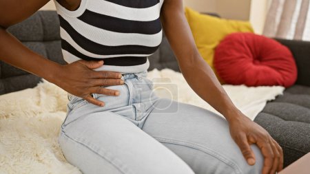 Téléchargez les photos : Belle adulte noire, une femme afro-américaine, souffrant d'un terrible mal d'estomac, assise sur un canapé à la maison, touchant son estomac douloureux, illustrant la douleur dans son portrait - en image libre de droit