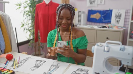 Foto de Mujer afroamericana con trenzas usando smartphone en tienda de costura rodeada de bocetos de moda y máquina de coser. - Imagen libre de derechos