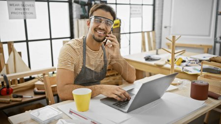 A smiling hispanic man with a beard in a woodworking workshop talks on the phone while using a laptop.