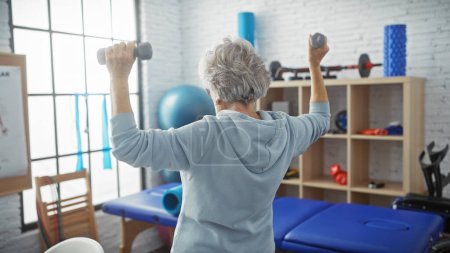 Photo for Senior woman exercises with dumbbells in a bright physiotherapy clinic showing fitness, health, and active aging. - Royalty Free Image