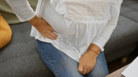 Senior woman sitting on couch indoors with hand on her hip in pain, depicting issues of health and aging.