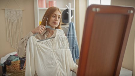 A beautiful, young, redhead woman evaluates a white blouse in a well-organized room with a mirror and fashionable decor.