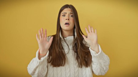 A surprised young hispanic woman with a white sweater poses against a yellow background, displaying a vivid emotional expression.