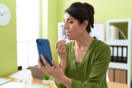 Photo for Middle age hispanic woman business worker using smartphone as a mirror make up lips at office - Royalty Free Image