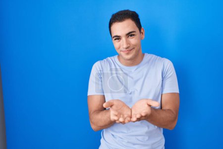Foto de Young hispanic man standing over blue background smiling with hands palms together receiving or giving gesture. hold and protection - Imagen libre de derechos