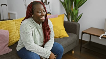 Téléchargez les photos : Femme africaine riante avec tresses rouges assise dans un salon moderne, respirant joie et confort. - en image libre de droit